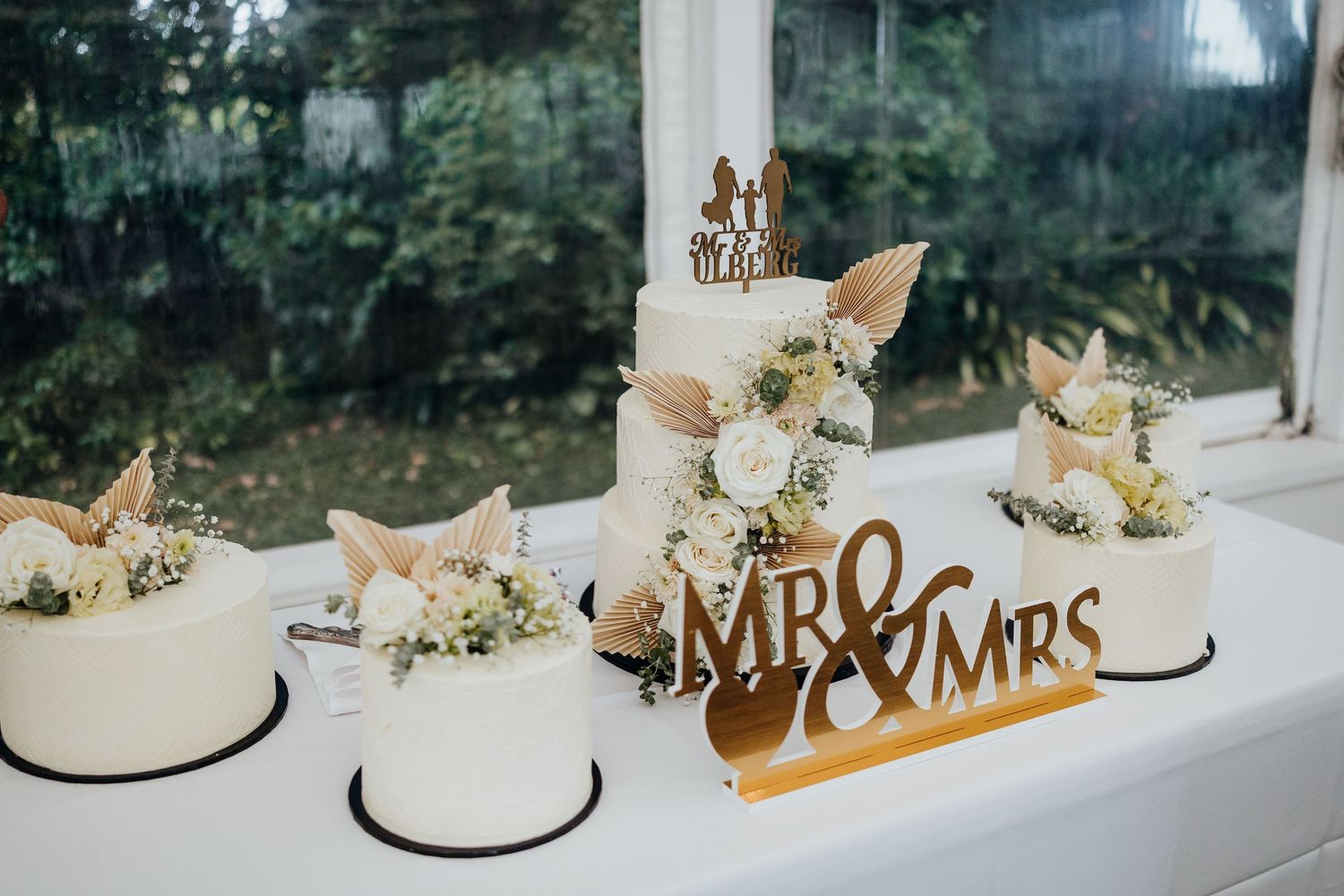 A white wedding cake decorated with gold accents and floral arrangements sits on display near a window.