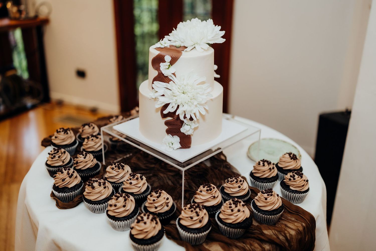A white wedding cake decorated with gold accents and floral arrangements sits on display near a window.