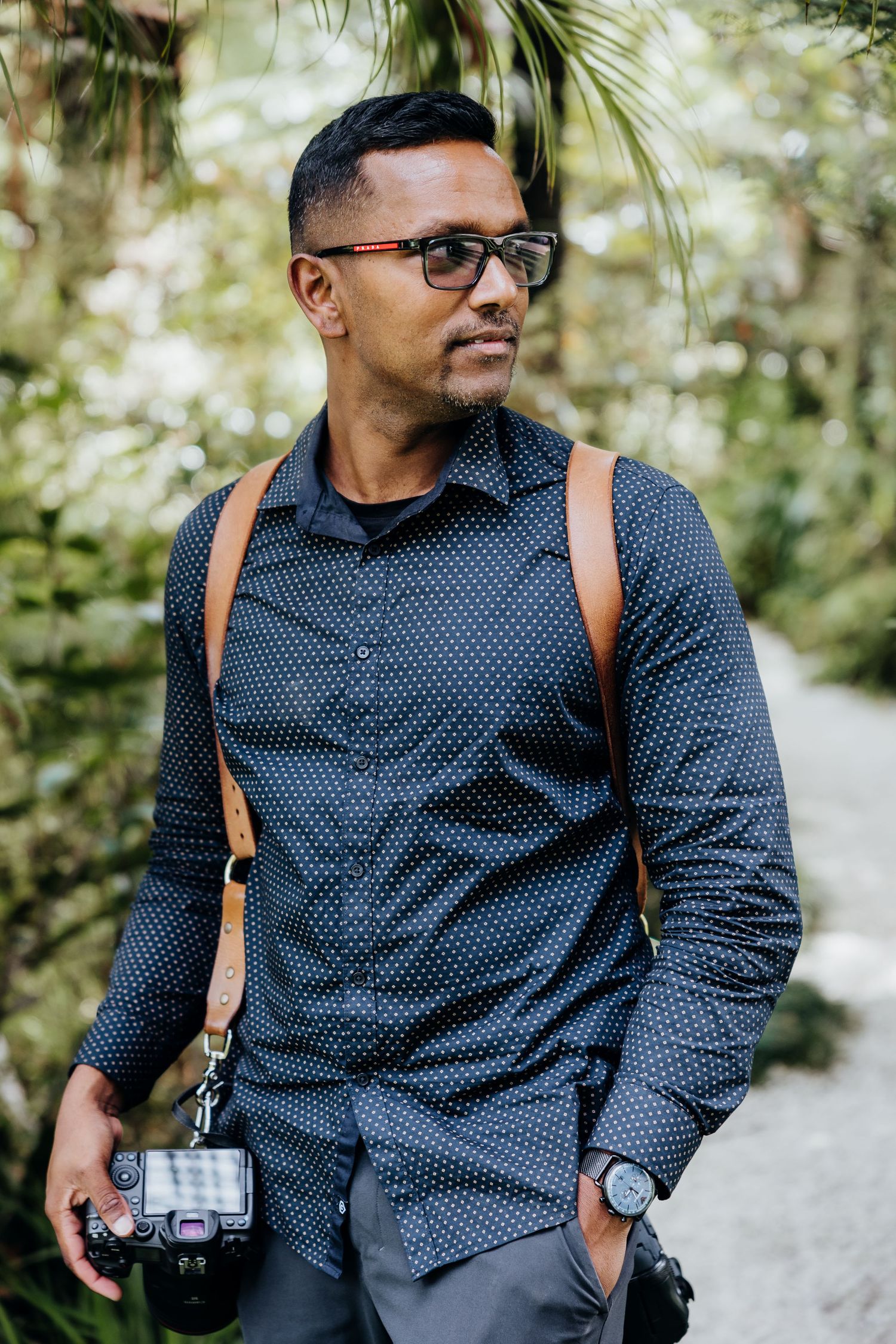 A person in a polka dot navy shirt and glasses poses with a camera in an outdoor nature setting.