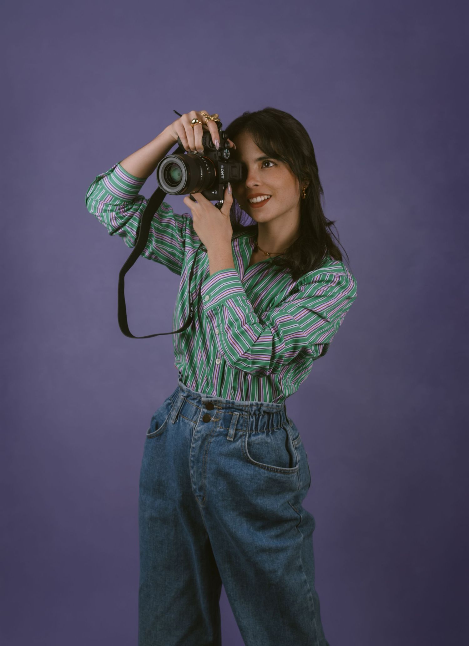 A photographer wearing a plaid shirt and jeans poses with a professional camera against a purple backdrop.