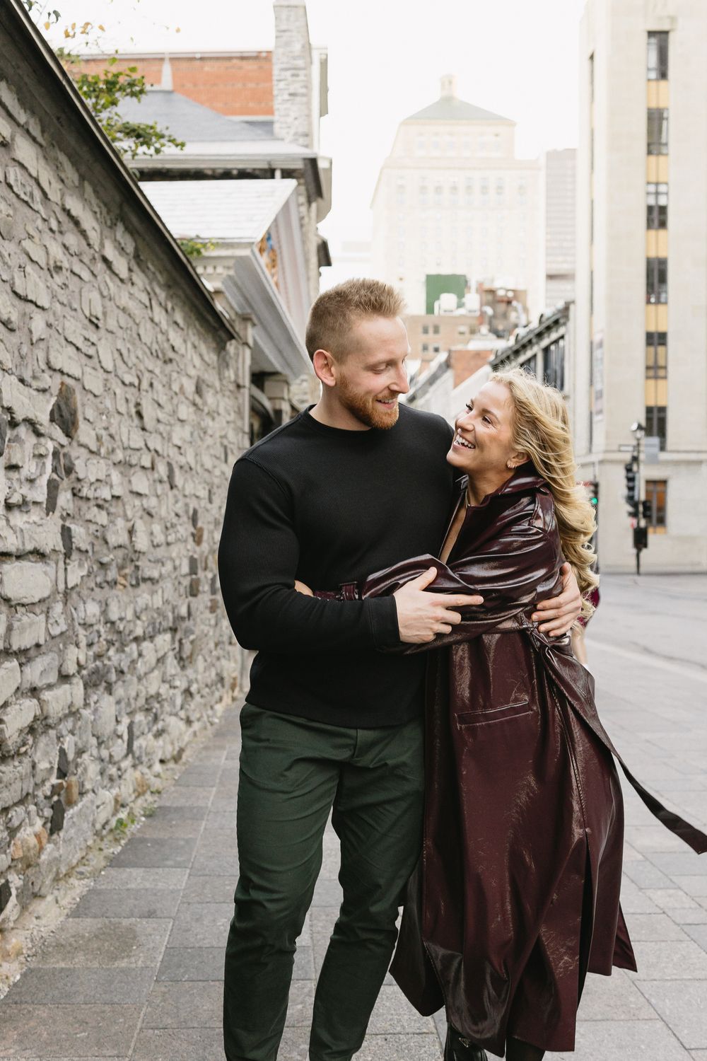 Happy couple laughing and embracing on a cobblestone city street beside a historic stone wall.