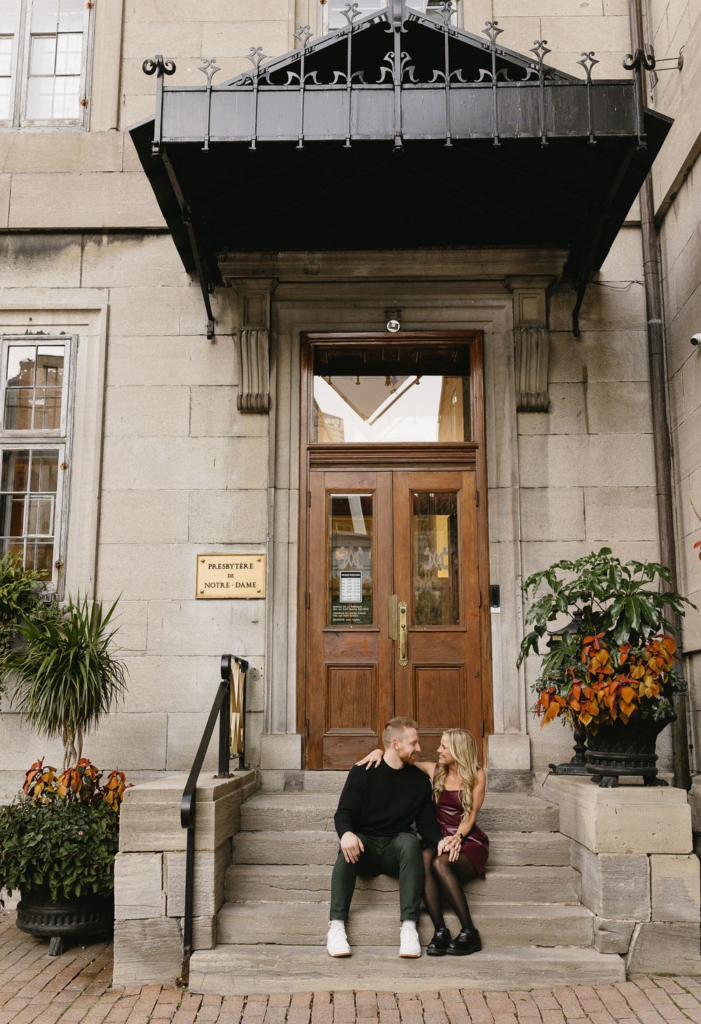 Smiling couple sitting on steps of elegant stone building with wooden doors and autumn decor.