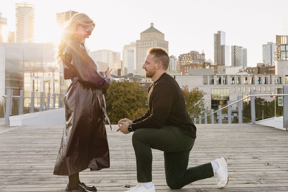 Man proposing on one knee to his partner on a rooftop deck with Montreal city skyline at sunset.