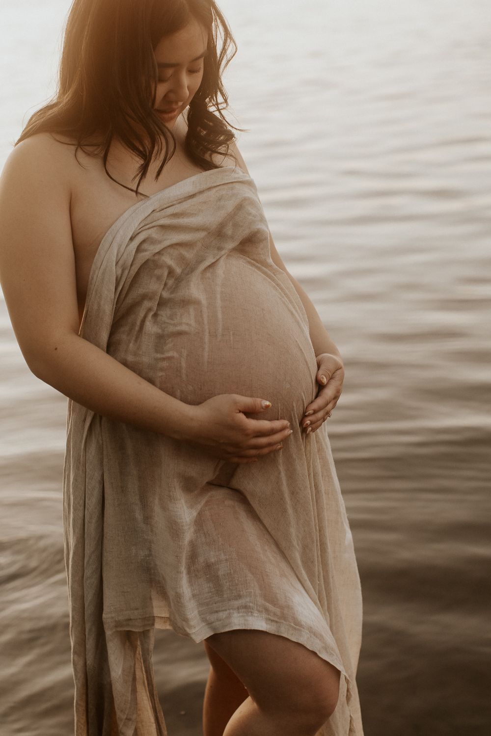 Pregnant woman wrapped in sheer linen fabric standing by water at golden hour, cradling her baby bump.