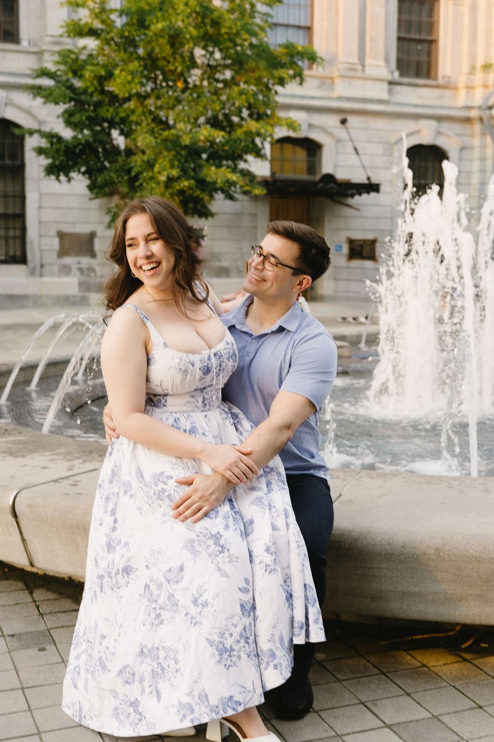 Smiling couple embracing by a fountain in an urban setting, woman in floral dress and man in blue polo shirt.