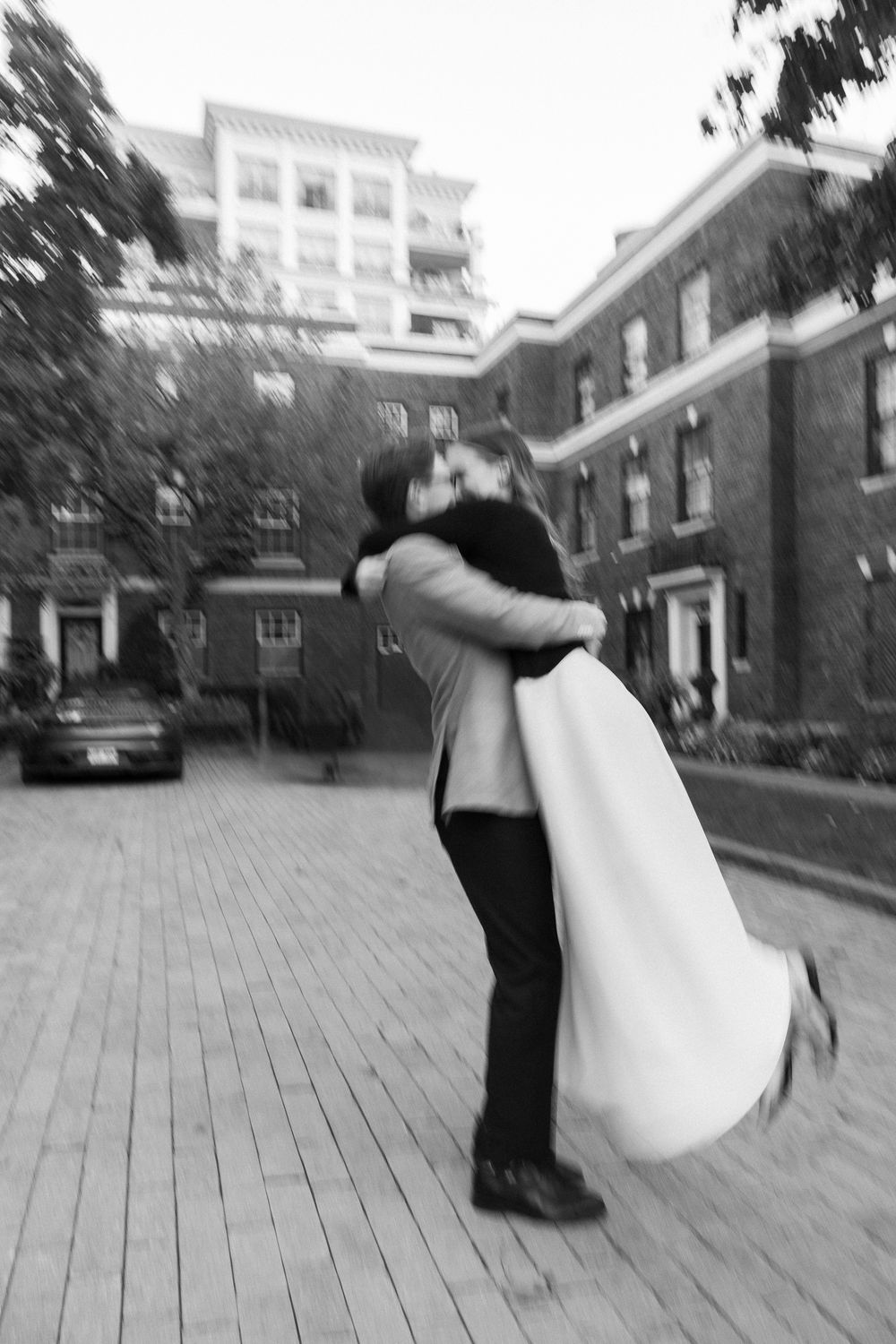 Black and white photo of groom lifting bride for a kiss outdoors on a wooden deck near brick buildings.