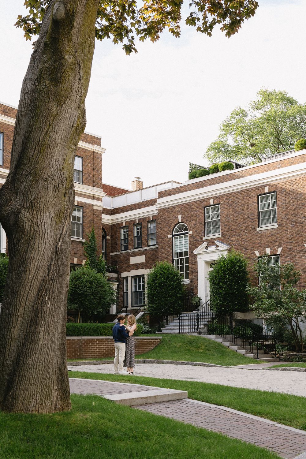 Engaged couple embraces near large tree outside classic brick building during outdoor engagement session in Montreal