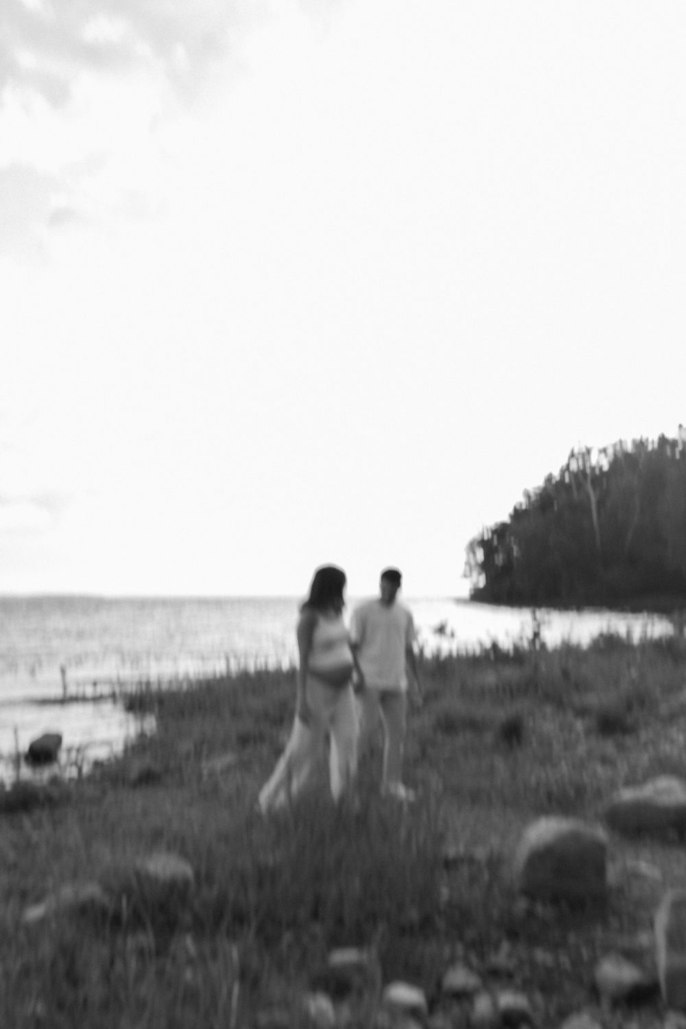 Black and white photo of a pregnant couple standing together near a serene lakeside landscape.