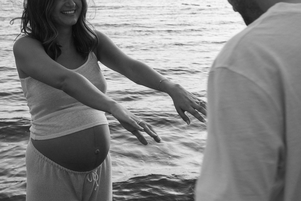 Black and white photo of a pregnant woman reaching toward her partner near the ocean waves.