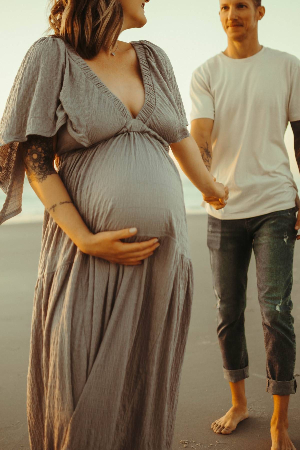 Maternity photoshoot at sunset on beach in flowing gray dress.