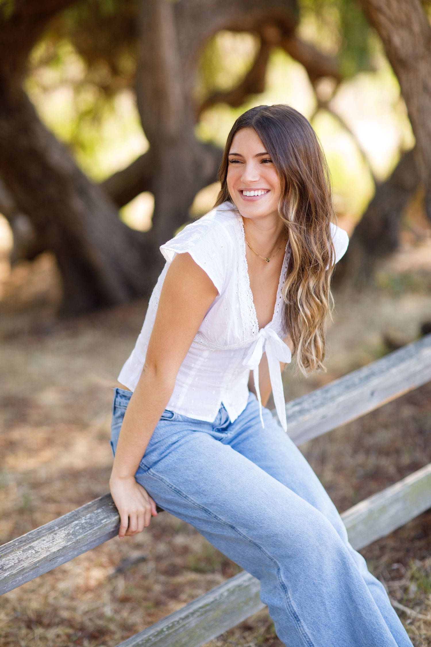 Casual outdoor portrait of a woman in a white tie-front top and jeans sitting on a rustic wooden fence.