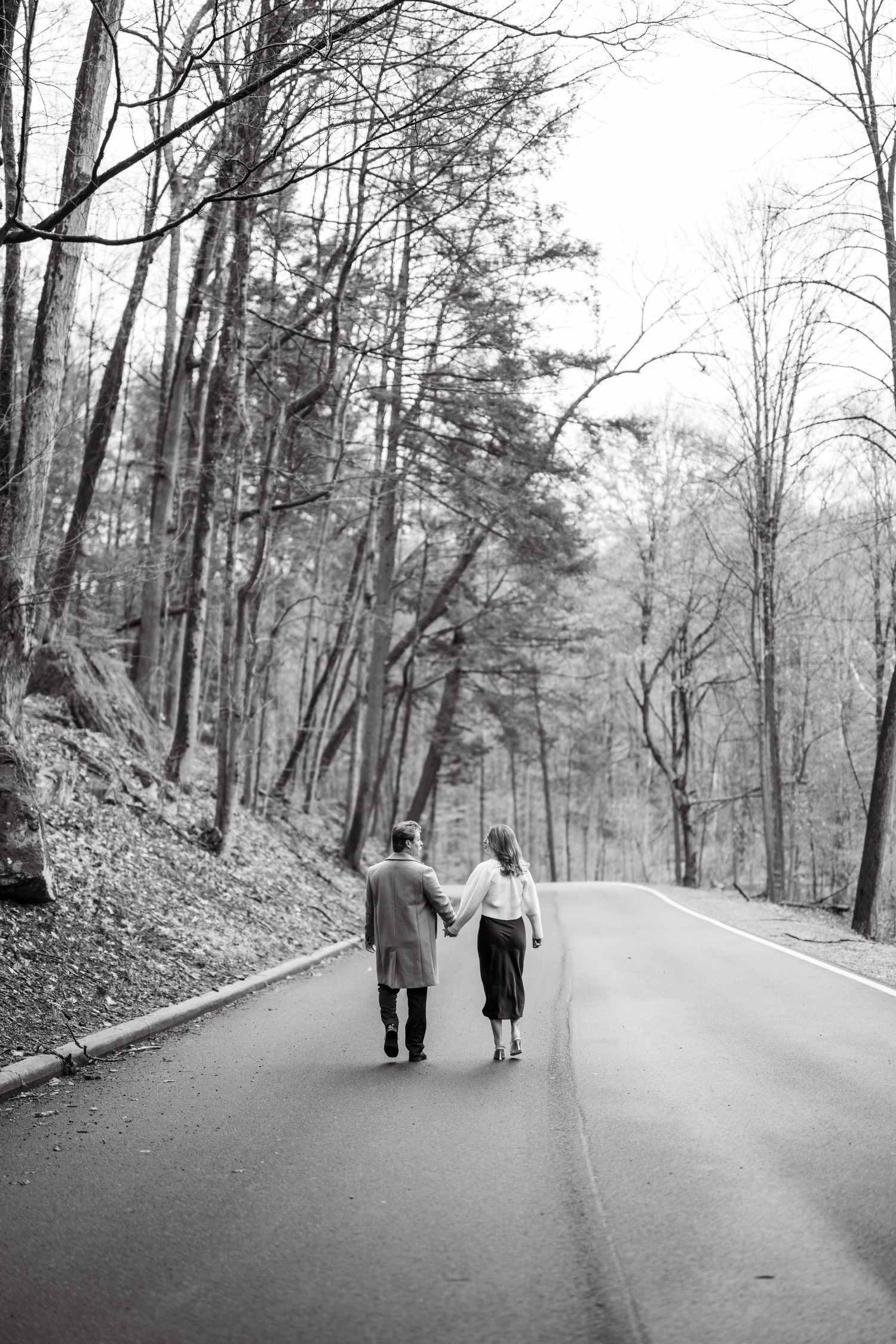 Two people walking together on an empty tree-lined road in a black and white vintage photograph taken from behind.