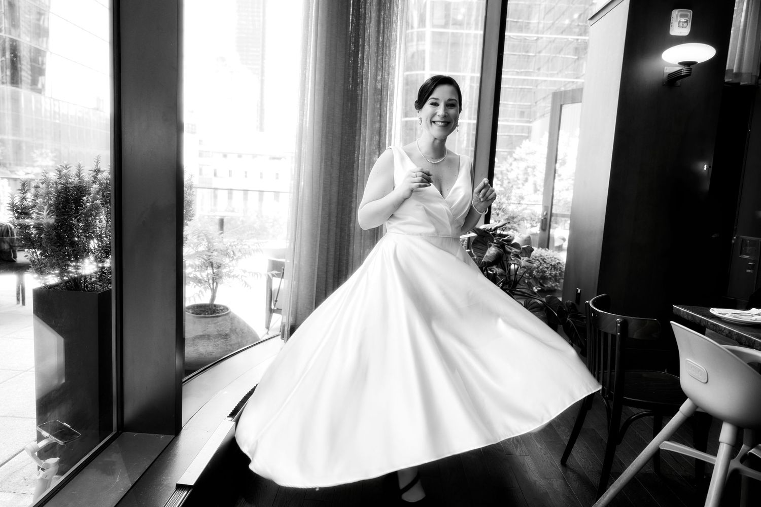 Bride in flowing white dress twirls joyfully in sunlit room with city views.
