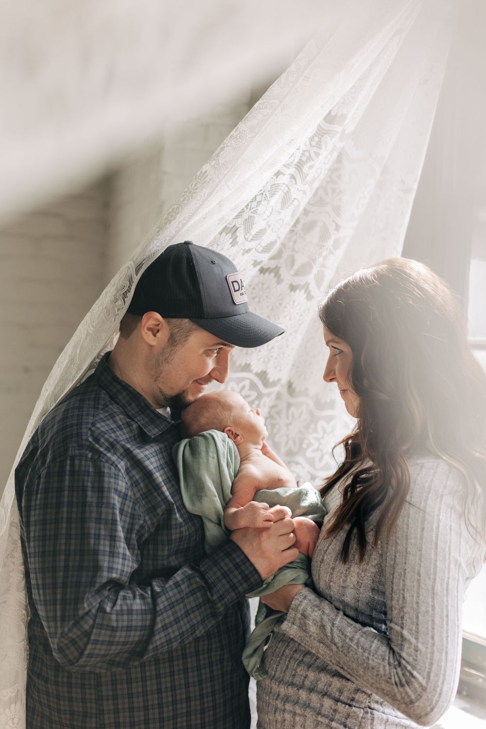Parents gazing lovingly at their newborn baby against a lace curtain backdrop.