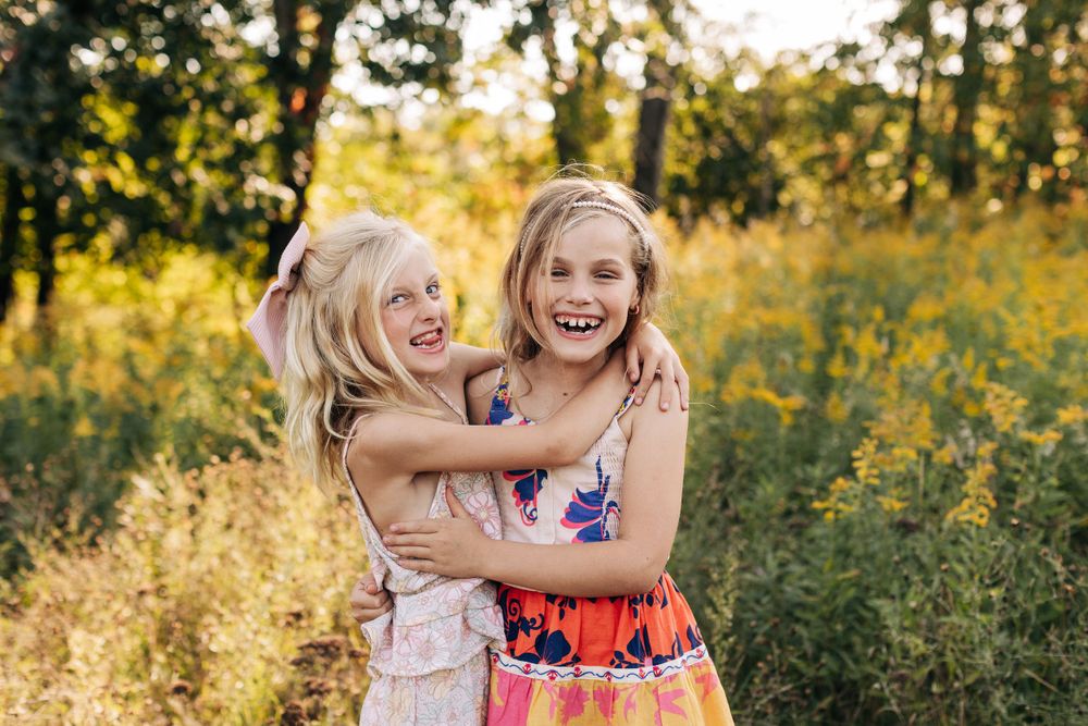 Two young friends share an enthusiastic embrace while laughing in a sunlit meadow.
