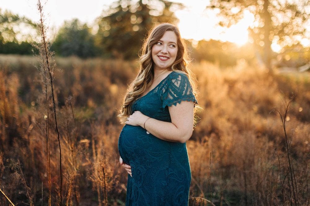 Expectant mother in teal dress poses during sunset in natural field setting.