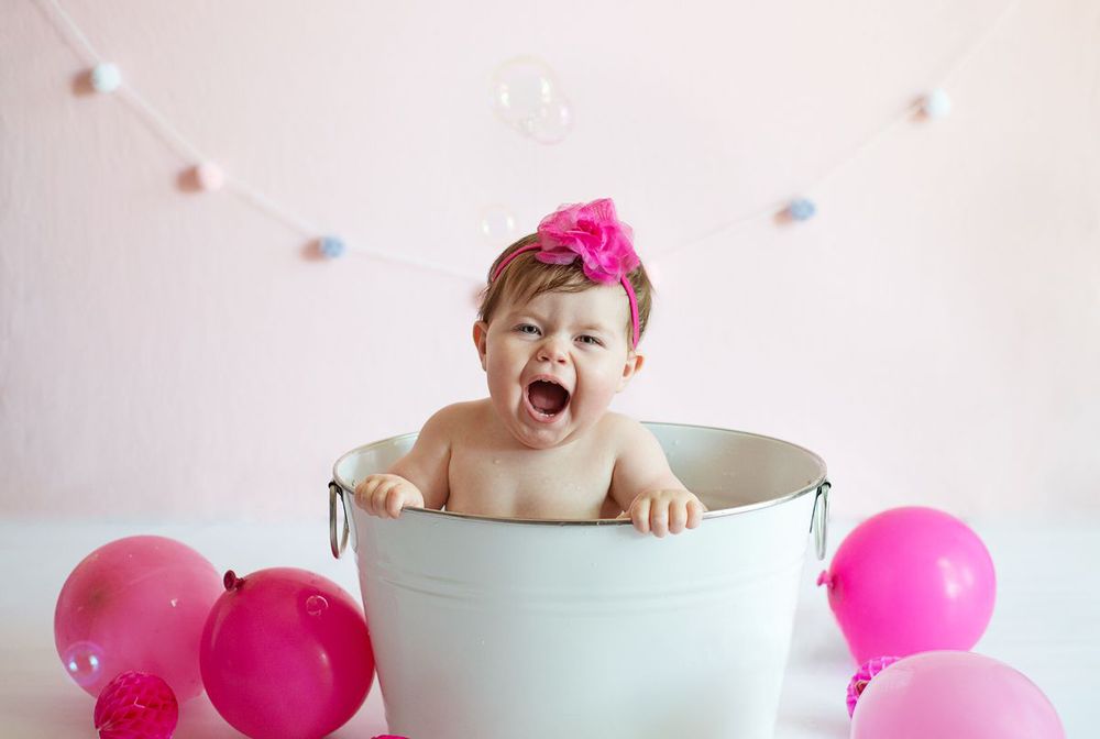happy one year girl gives a big smile while in a little white tub looking at the camera with pink balloons
