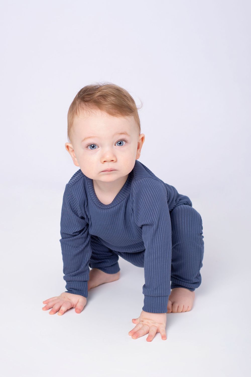 high key image of one year old in blue cotton outfit crouches down looking at camera