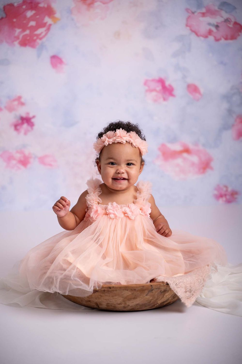 baby girls smiles with first birthday in wood prop wearing pink tulle dress with floral backdrop for smash cake photos in studio, Cobourg*
