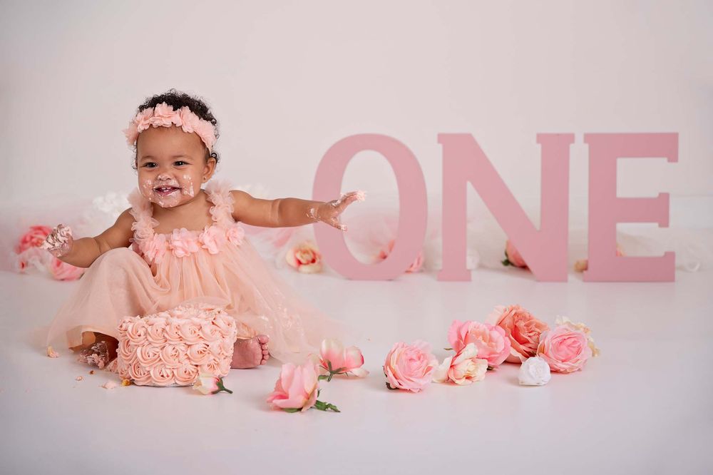 baby girls smiles with first birthday cake on her face wearing pink tulle dress surrounded in pink roses and smash cake in studio, Cobourg*