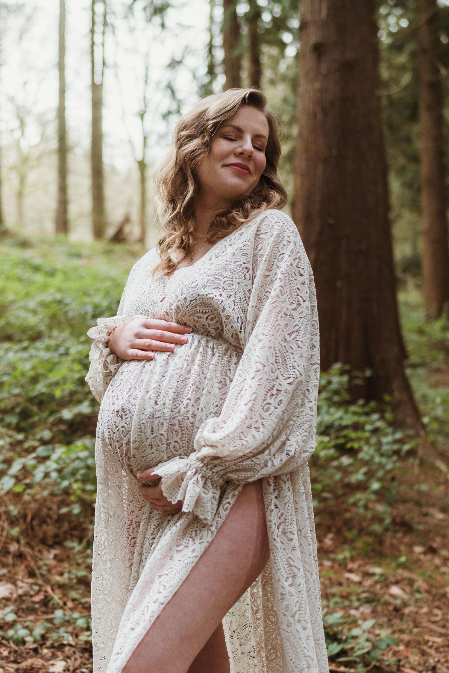 Maternity photo of woman in white lace dress posing in forest setting.