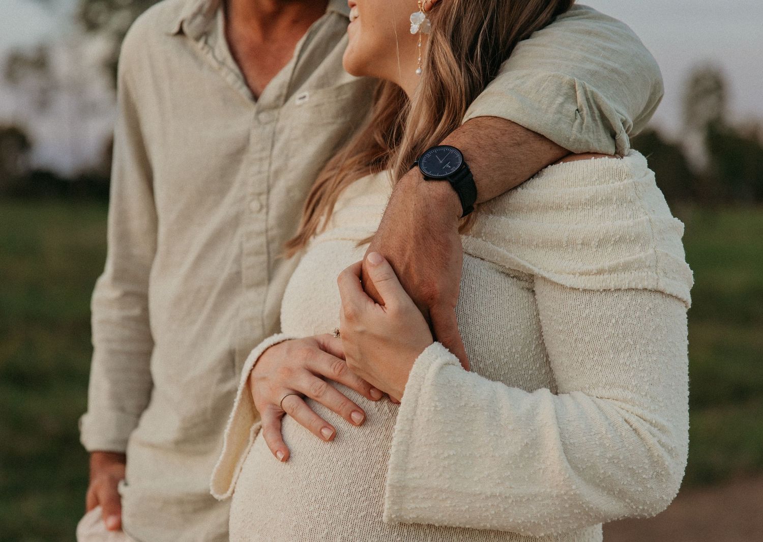 Woman hugging man from behind piggyback style, both laughing outdoors near water at golden hour.