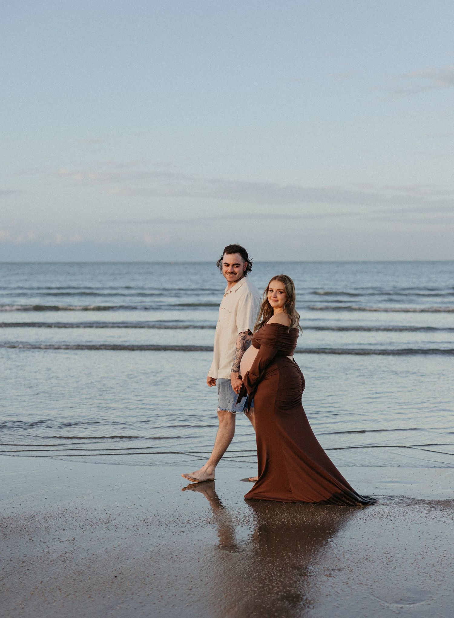 Pregnant couple walking on beach at sunset, woman in flowing brown gown, man in white shirt and denim shorts.