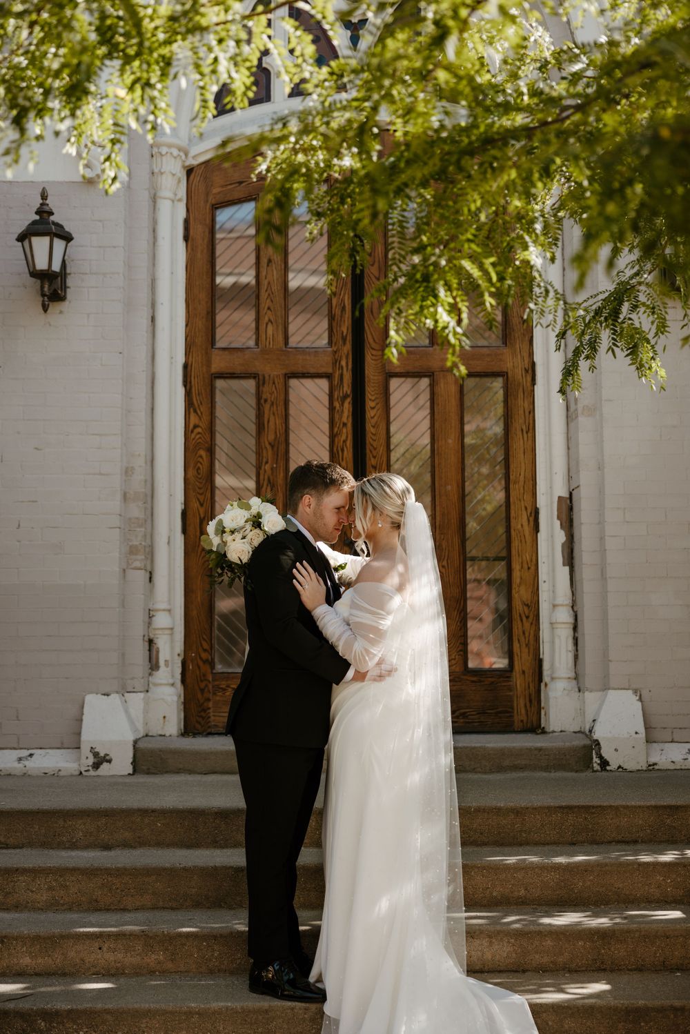 A bride and groom embrace on the steps of a building with a wooden door and hanging plants. Fort Worth Texas wedding photographer.