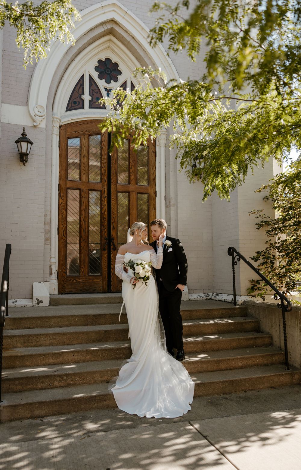 A bride and groom stand on the steps of a grand, ornate church building with arched doorways and lush greenery surrounding the entrance.