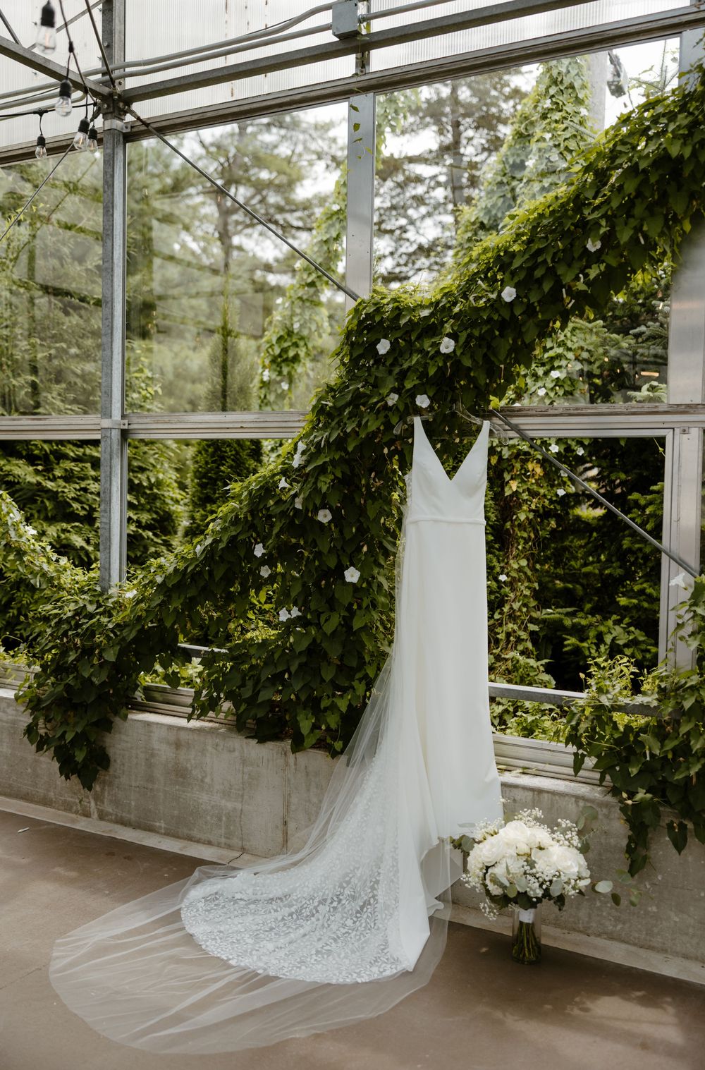 A white lace wedding dress with a long train hangs in a greenhouse surrounded by lush greenery. Fort Worth Texas wedding photographer.