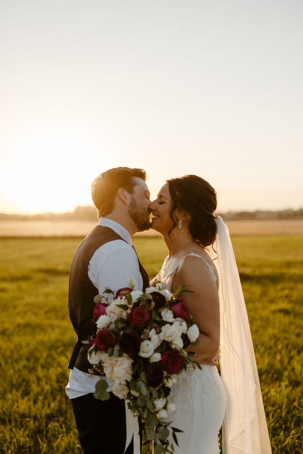 A bride and groom embrace in a field at sunset, holding a bouquet of flowers. Fort Worth Texas wedding photographer.