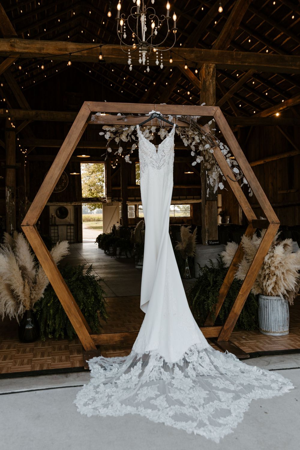 A white lace wedding dress with a long train hangs in a rustic wooden frame inside a dimly lit barn with a chandelier overhead. Fort Worth
