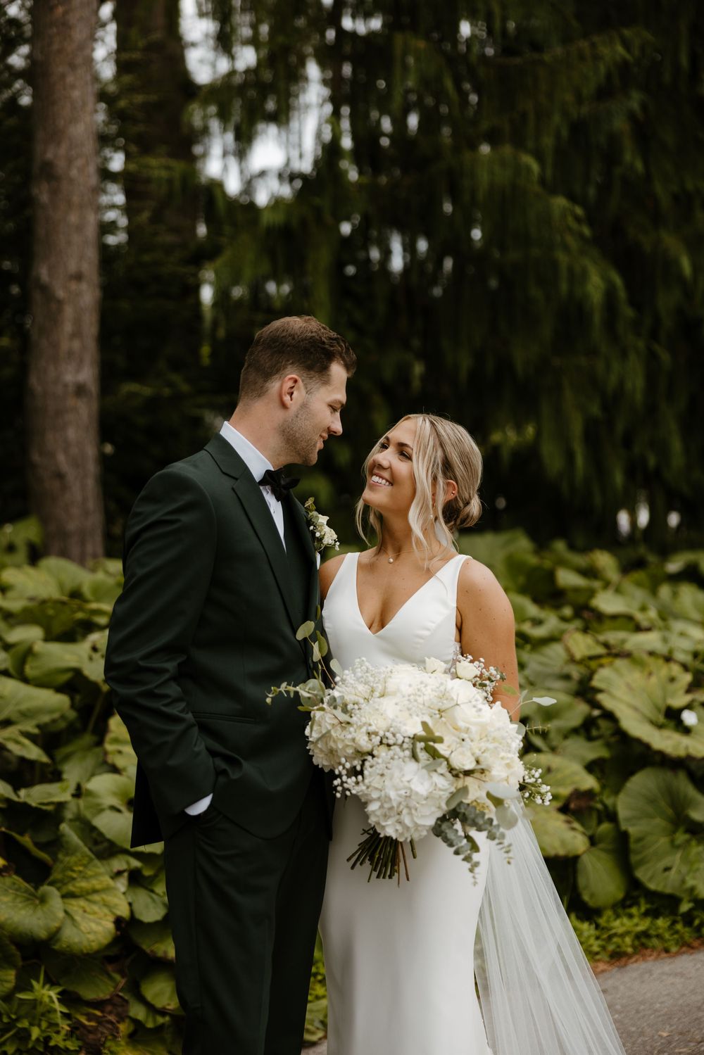 A bride and groom stand together in a lush, green outdoor setting, the bride holding a bouquet of white flowers. Fort Worth Texas