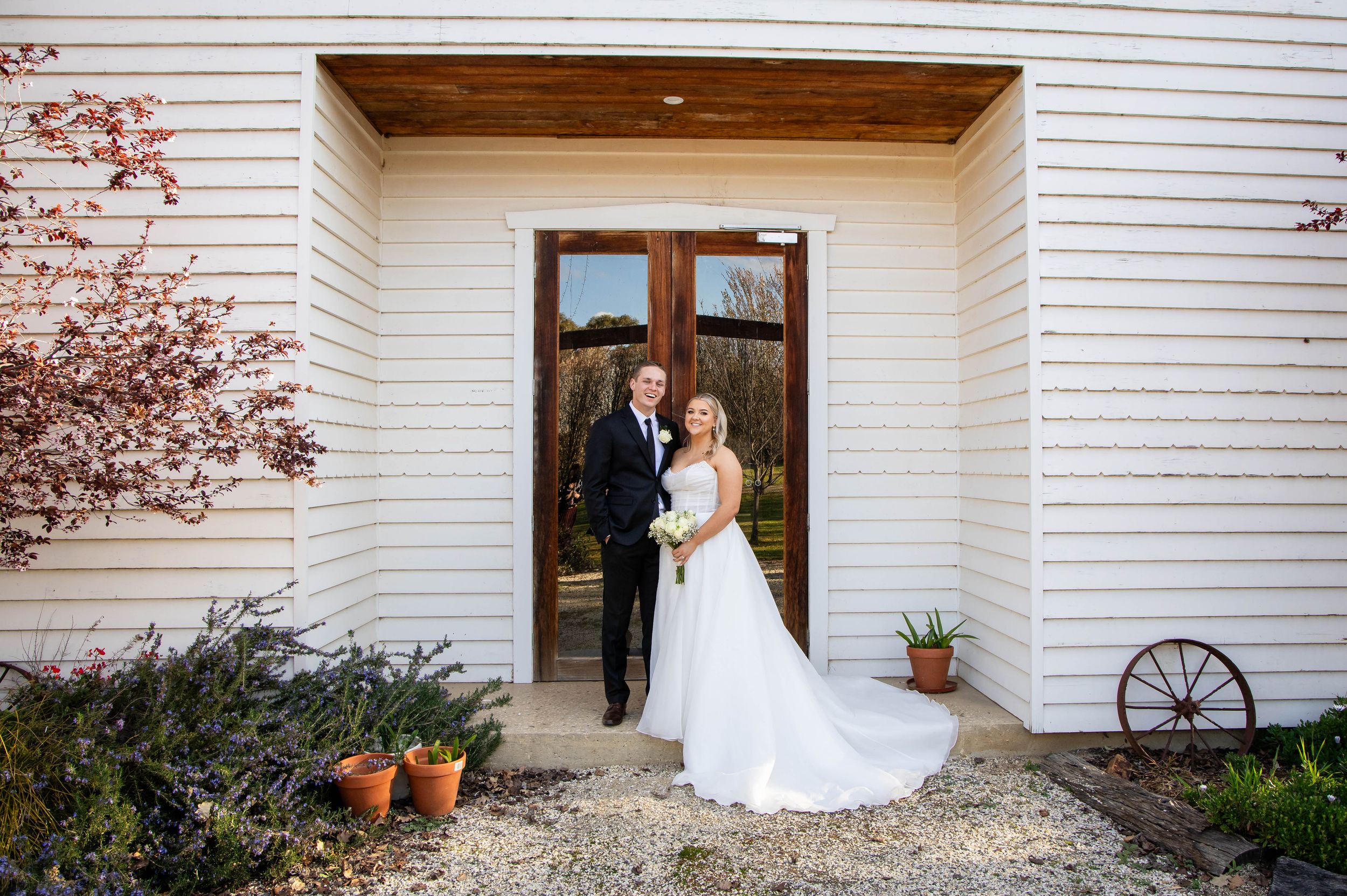 A couple stands in the doorway of a white siding building in formal wedding attire for a celebratory portrait.