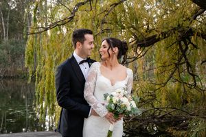 A couple shares an intimate moment under a weeping willow tree beside a pond while holding a white flower bouquet.