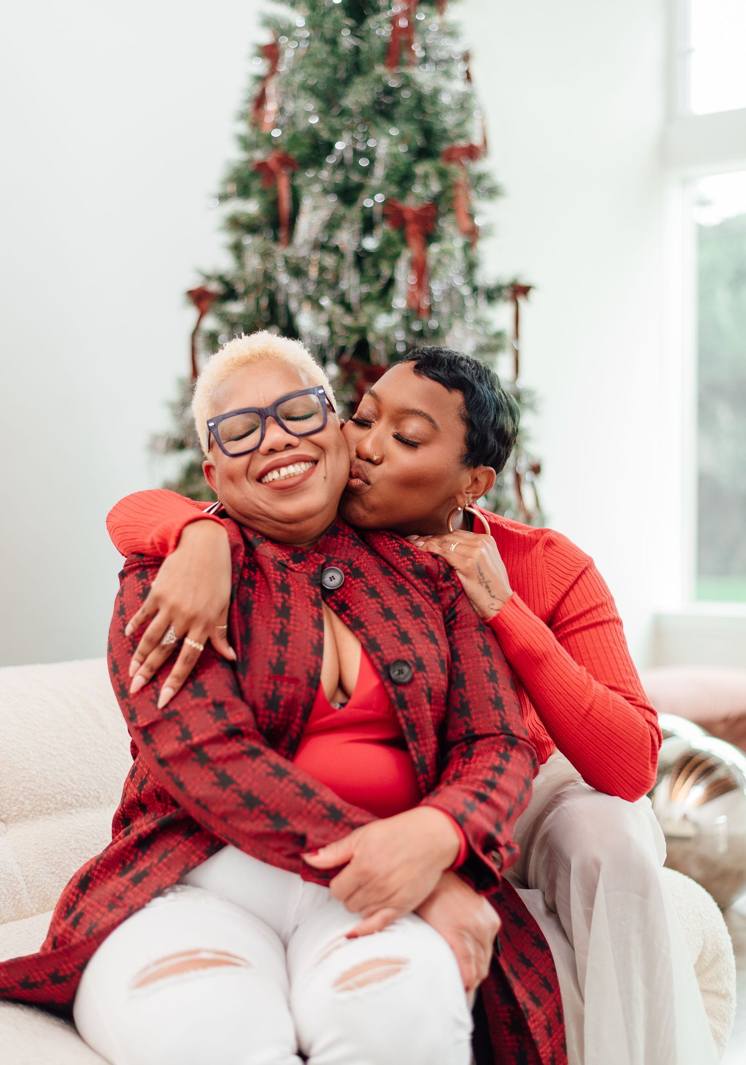 A couple in matching red holiday outfits share loving embraces and laughter in front of a decorated Christmas tree.