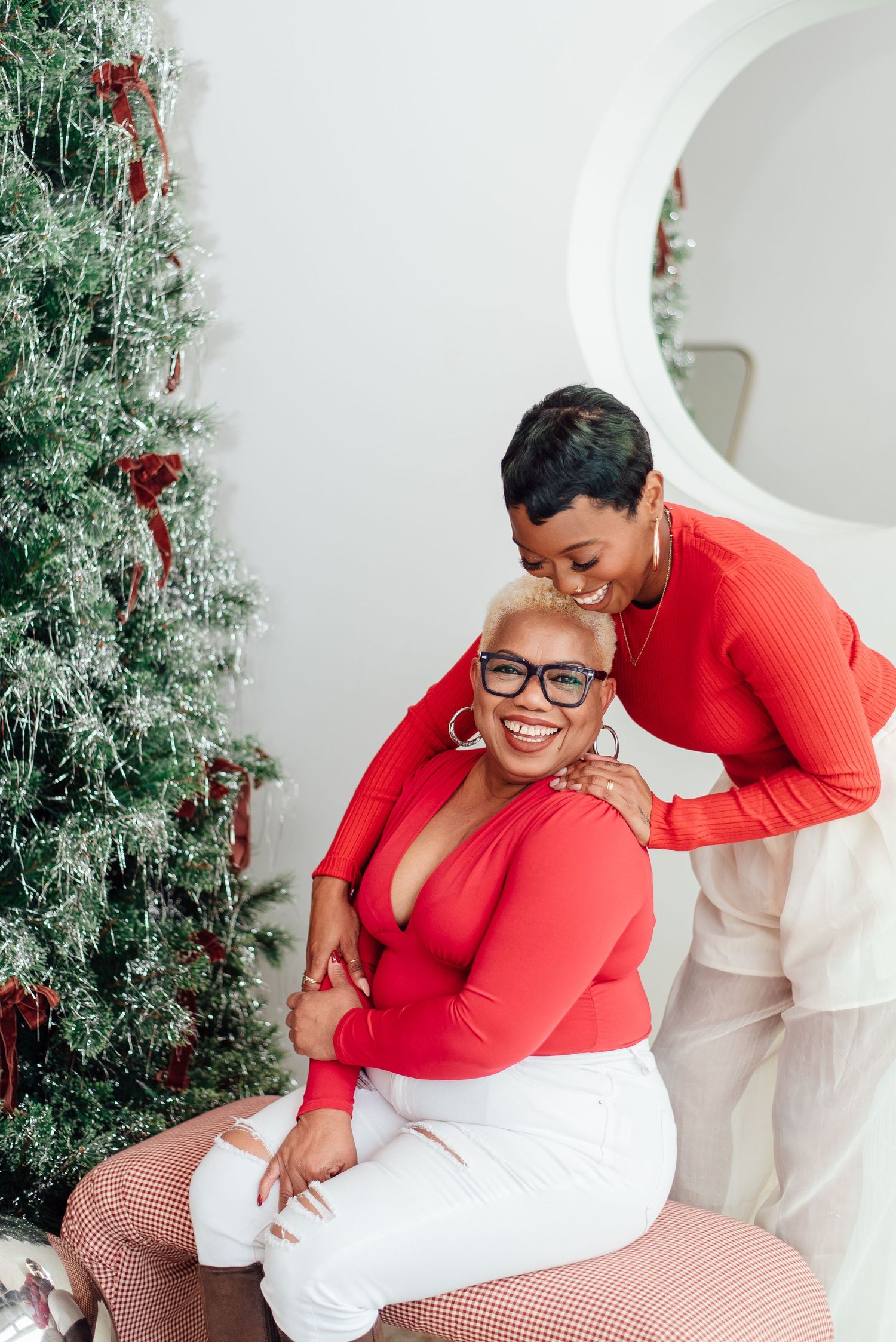 Two people wearing matching red tops share a joyful moment by a decorated Christmas tree in a bright white room.