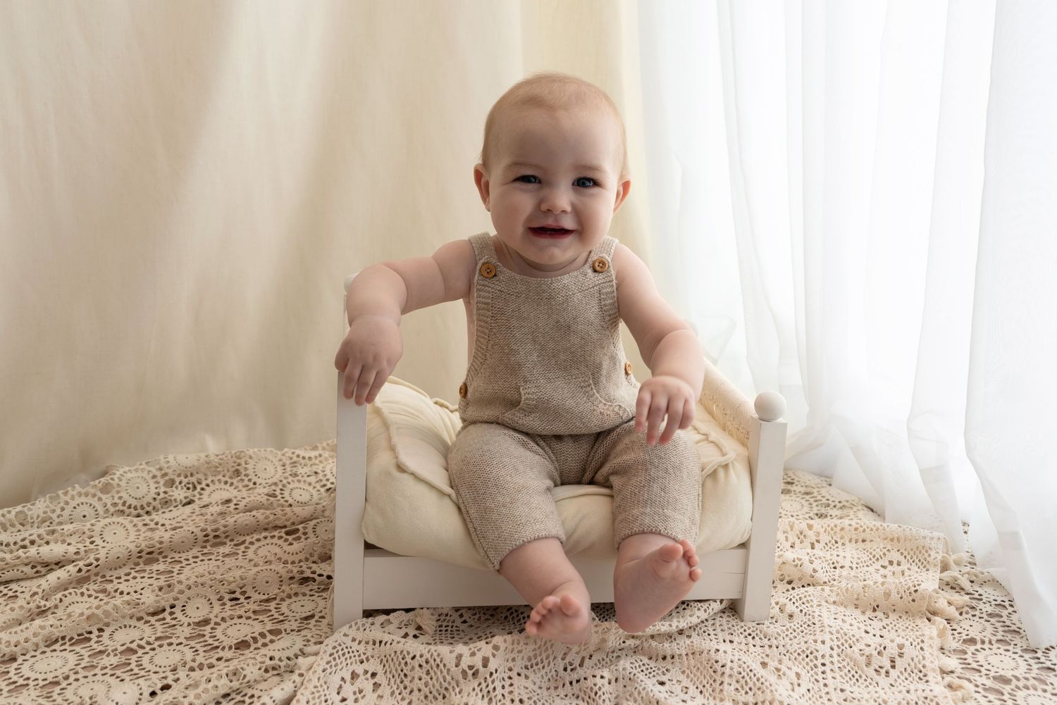 Smiling baby sits in small white chair wearing beige outfit on textured neutral carpet near sheer curtains.