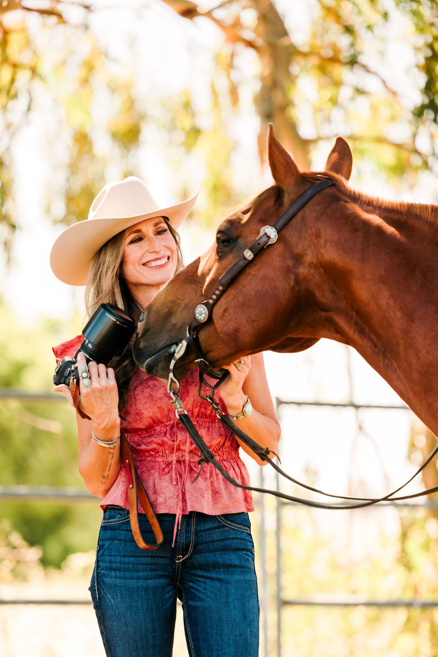 Photographer in western attire and cowboy hat with camera equipment poses alongside bay horse during golden hour.