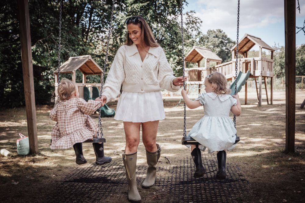 Mother holding hands with two young daughters at a sunny playground, wearing white outfit and knee-high boots.