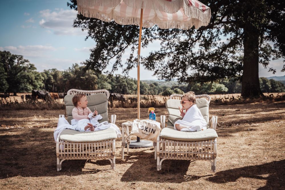 Two young children relaxing on white wicker lounge chairs under a boho umbrella in a sunny outdoor setting.