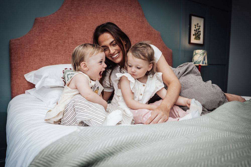 Smiling mother cuddling two toddler daughters on a bed with pink headboard in a cozy bedroom.