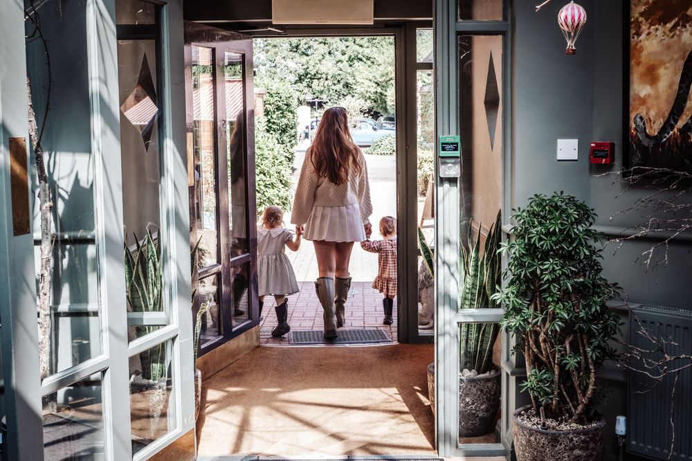 Woman holding hands with two young children walking through a doorway into bright sunlight.