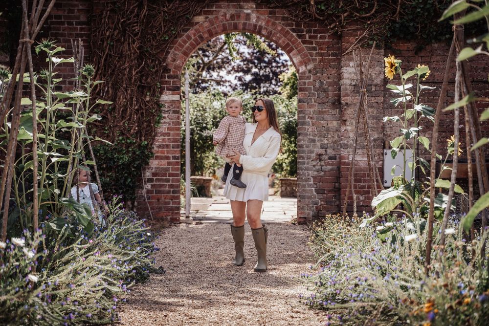 Woman in white dress holding baby in lush garden with brick archway and wildflowers.