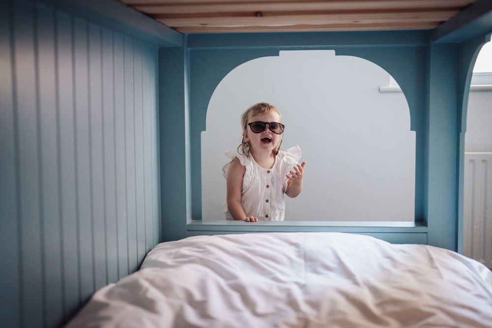 Happy toddler wearing sunglasses standing in a blue wooden bunk bed frame holding a small object.