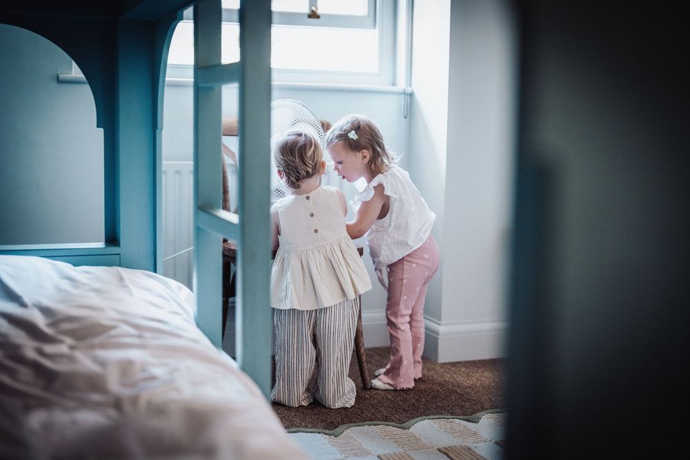 Two young girls in a blue bedroom, one helping the other dress in striped pants and white top.