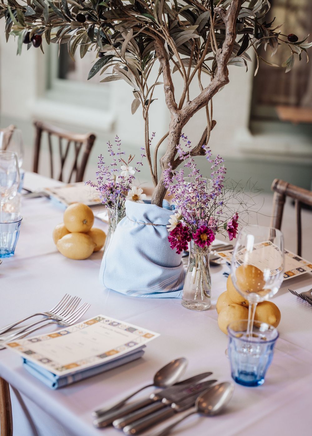 Elegant dining table set with lemons, wildflowers, blue glassware, and an olive tree centerpiece.