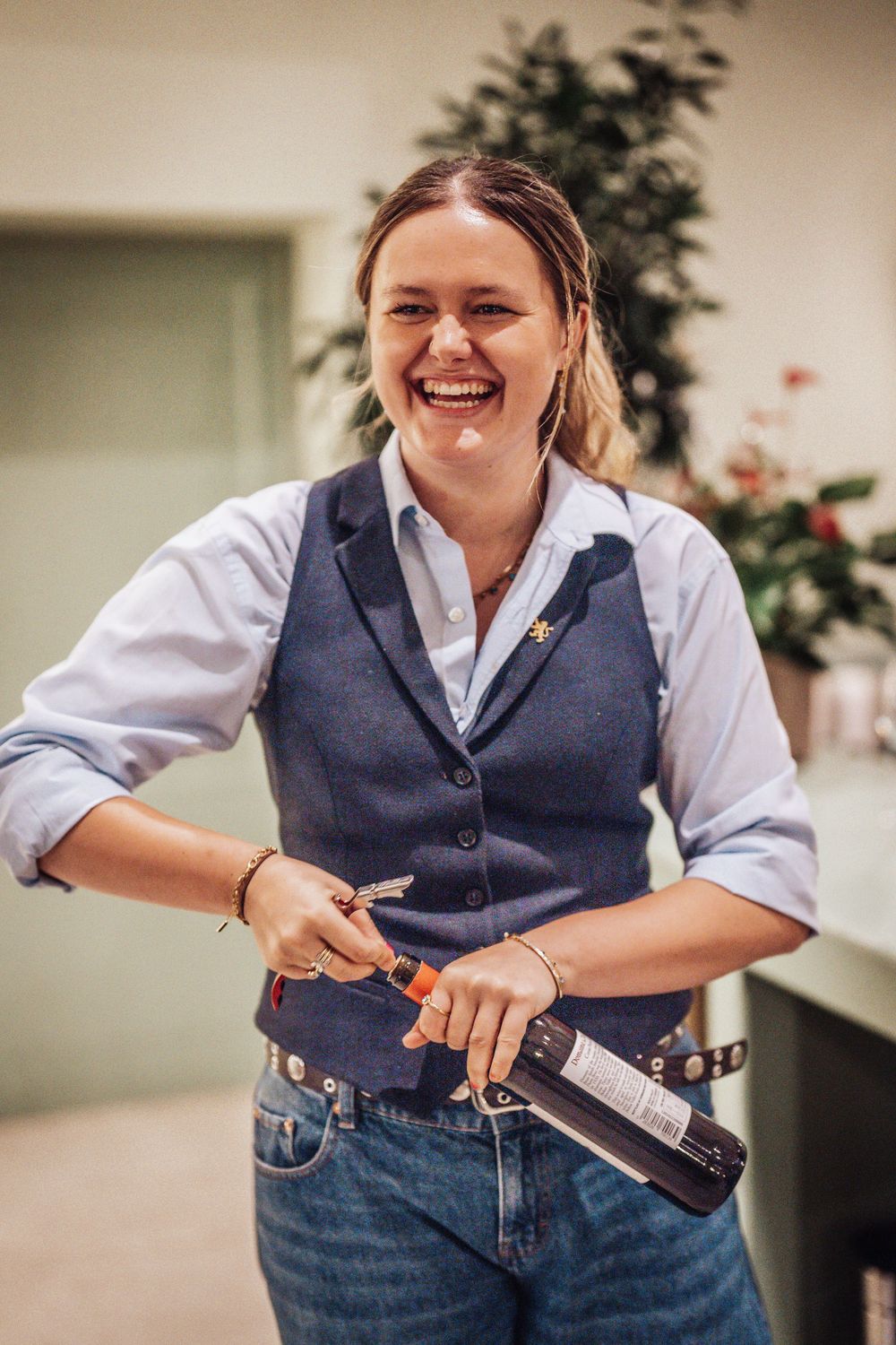 Smiling female server laughing while opening a wine bottle wearing a blue vest and white shirt.