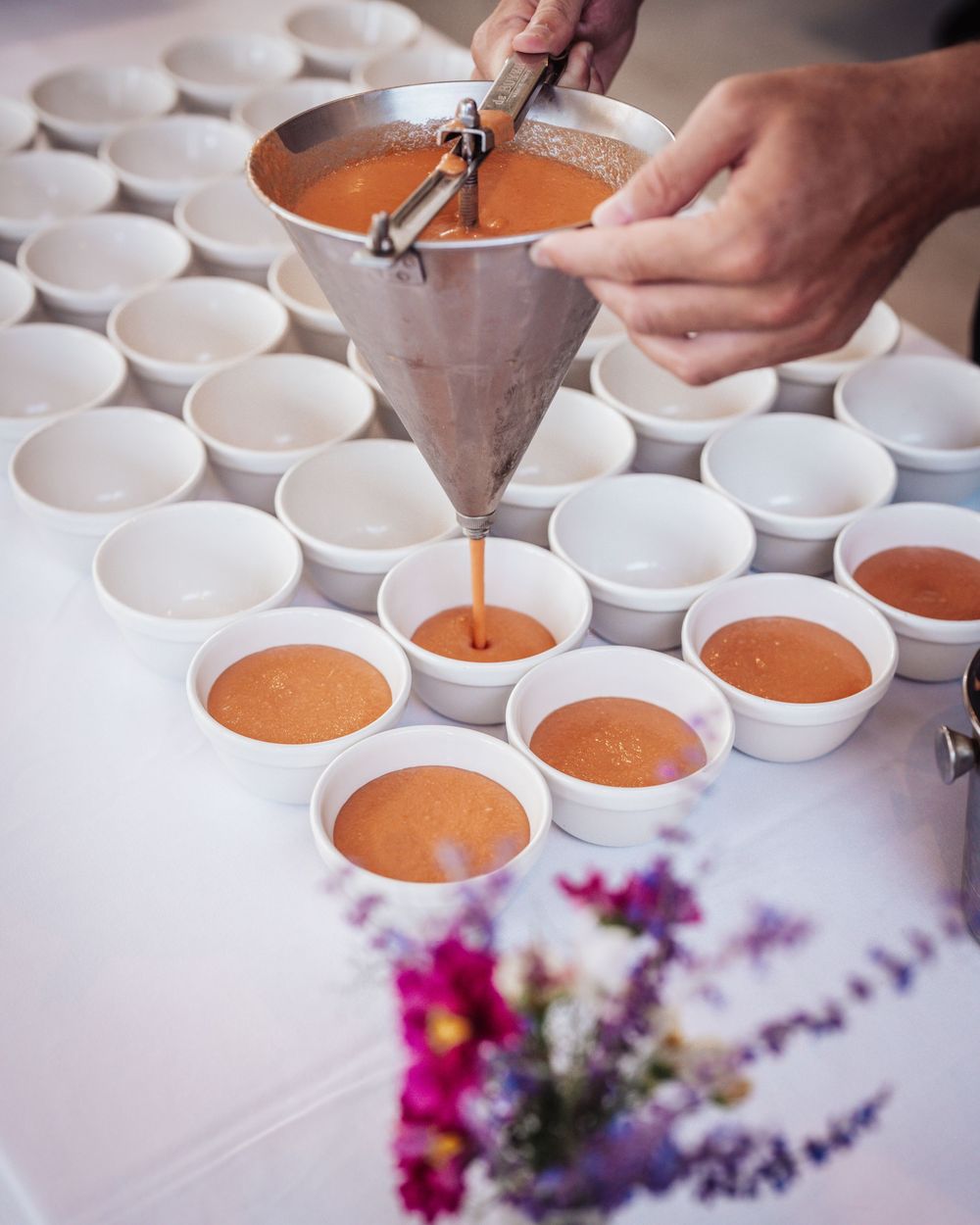 Chef pouring orange tomato soup bisque into white ramekins using a funnel at a catering event.
