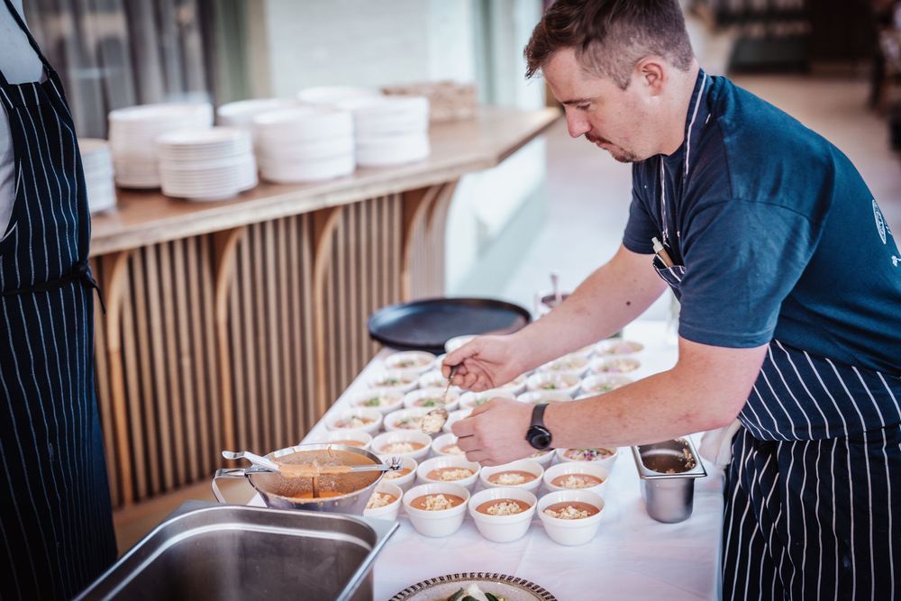 Chef in apron carefully plating individual dessert cups on a catering table in a modern kitchen setting.